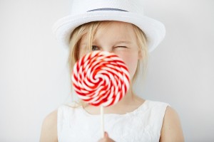 Close up portrait of beautiful little female child in white hat holding huge spiral lollipop having fun while enjoying sweet candy with happy expression against white concrete wall. Selective focus